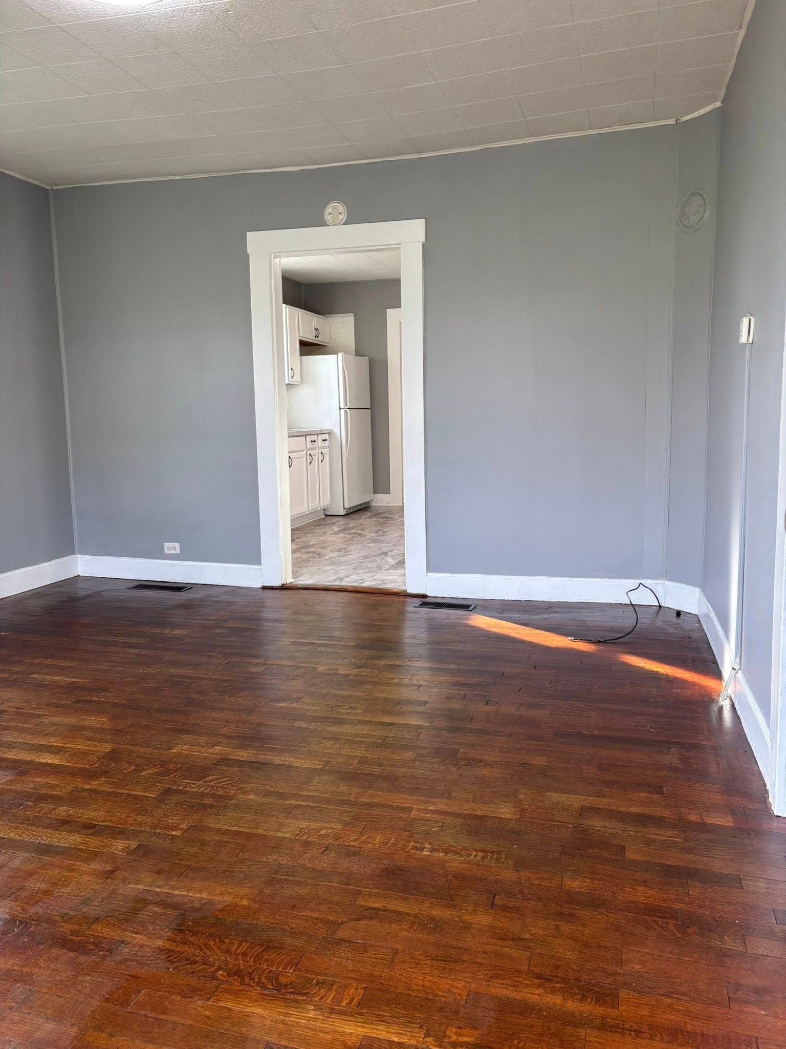 Empty room with gray walls, white trim, and stained wood floor; doorway to kitchen.