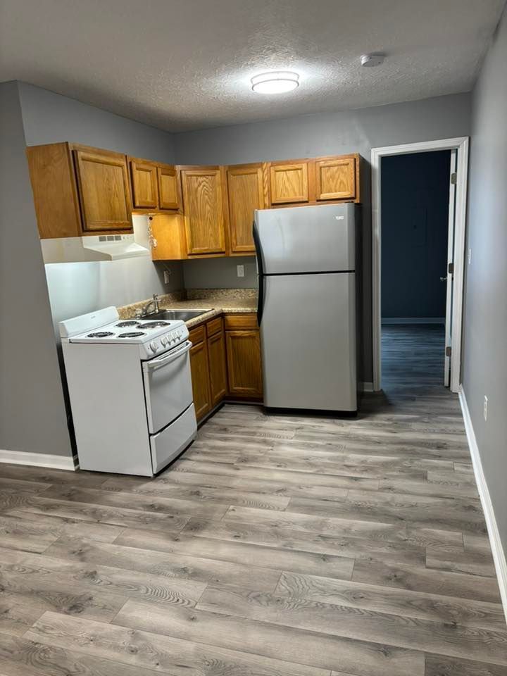 Small kitchen with wooden cabinets, white appliances, and gray flooring. A door leads to another room.