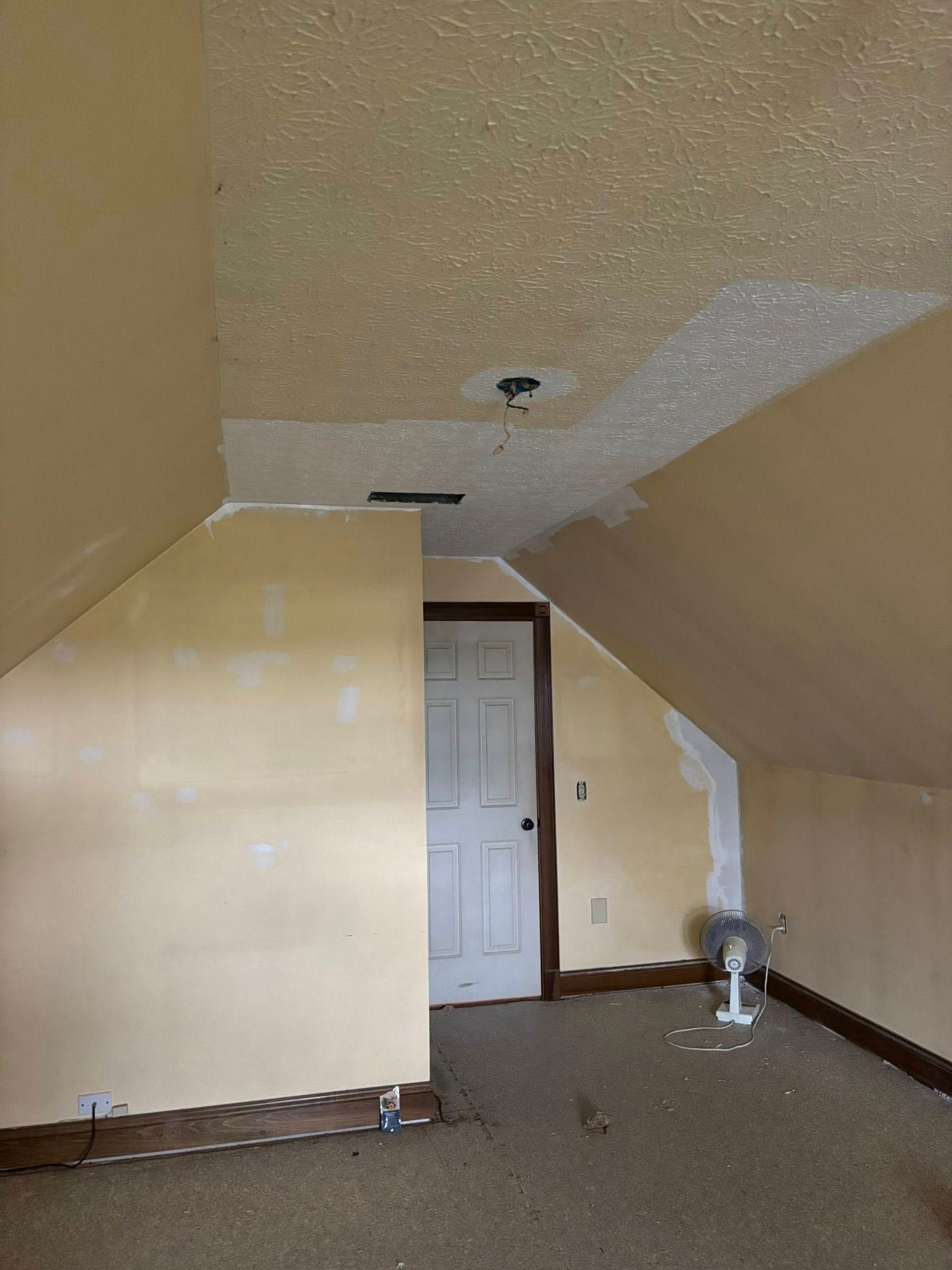 Room with sloped ceiling, door, and a fan; walls and ceiling are different shades of beige.