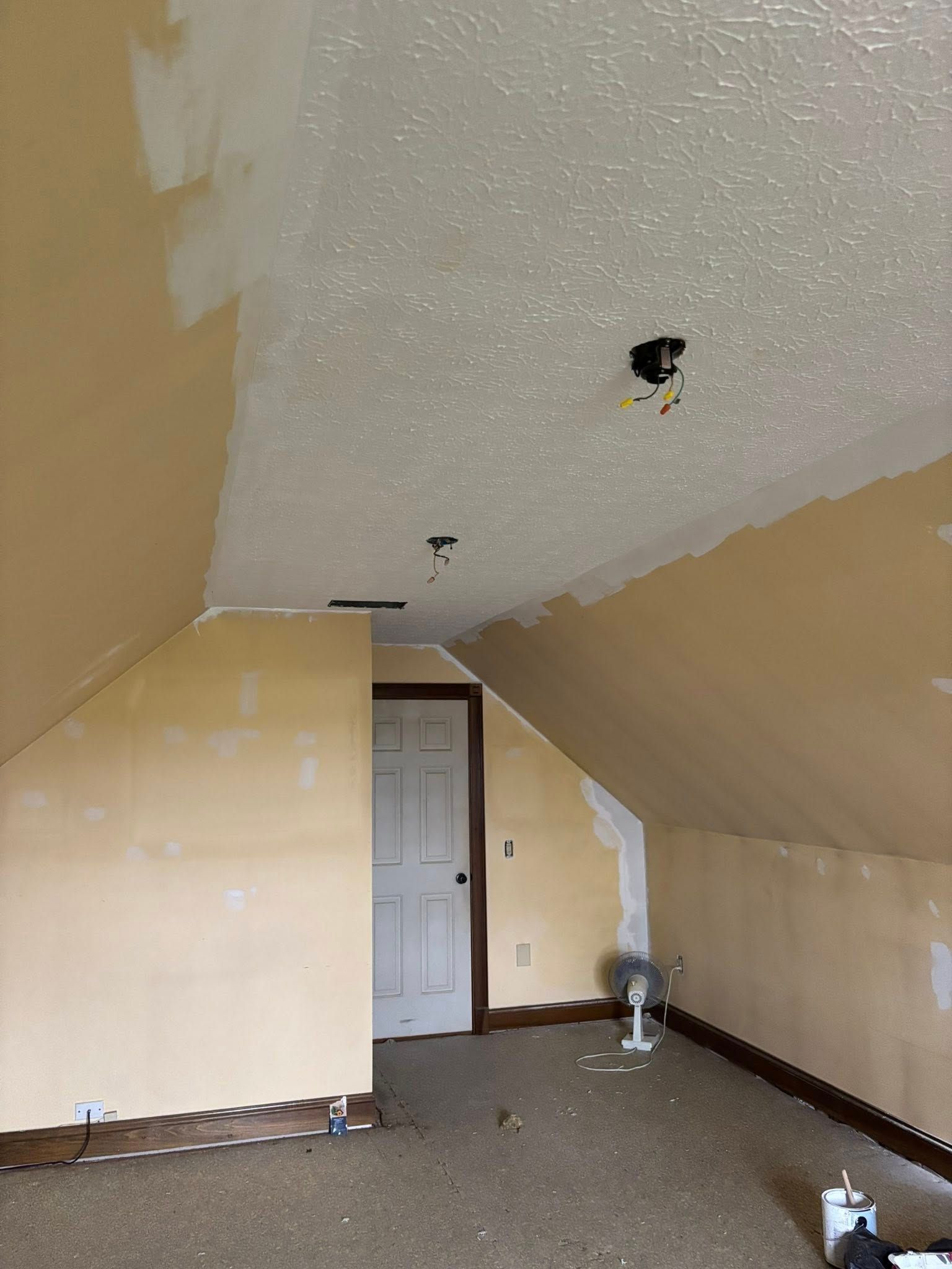 Attic room with sloped walls, painted light yellow with textured ceiling. Door visible, fan on floor.