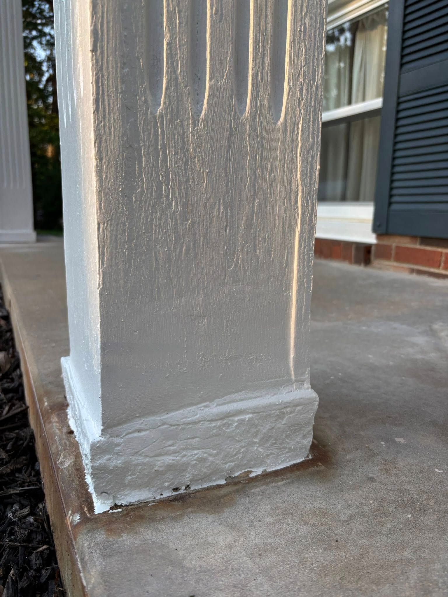 Gray, fluted square porch column with dripping paint. Stands on a concrete porch, next to window and a dark shutter.