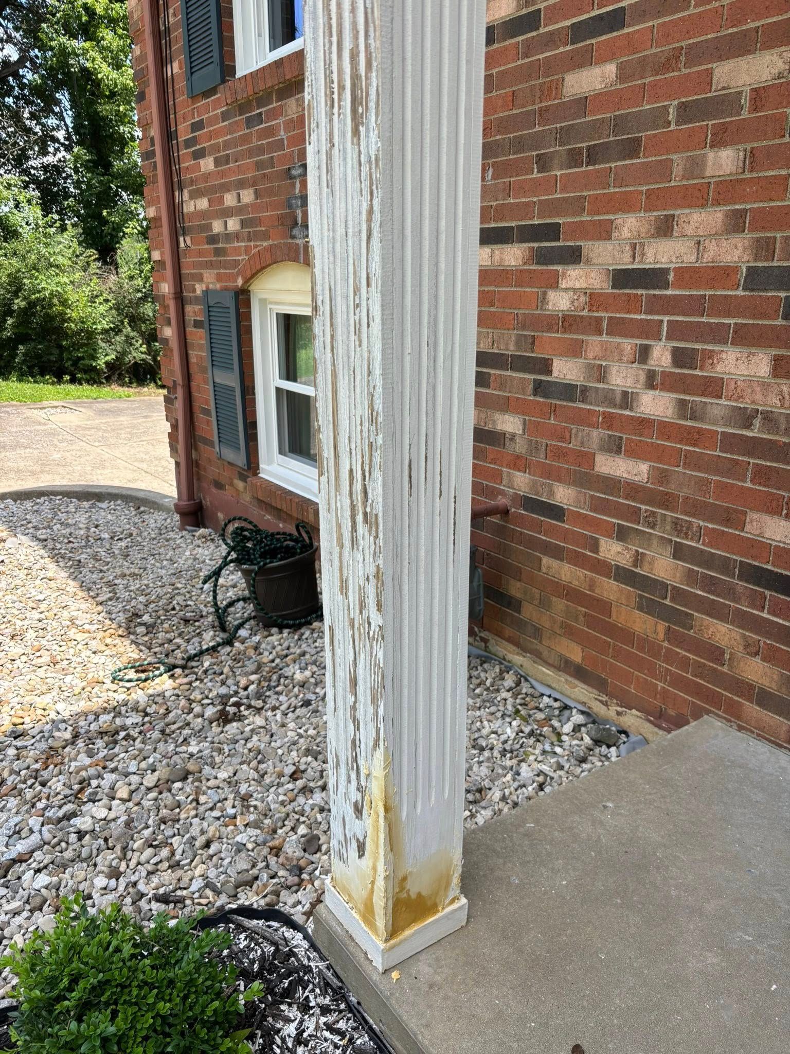 White column with peeling paint next to brick house. Gray stone ground and small bush in the foreground.
