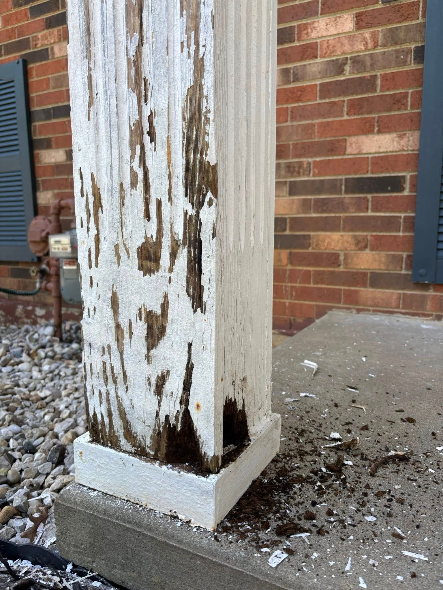 Damaged, peeling paint on a white pillar next to a brick wall. Debris on the ground.
