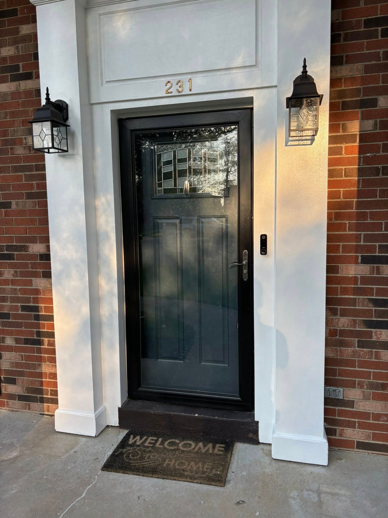 Front door with black frame, address 231 above, two sconce lights, brick exterior, welcome mat.