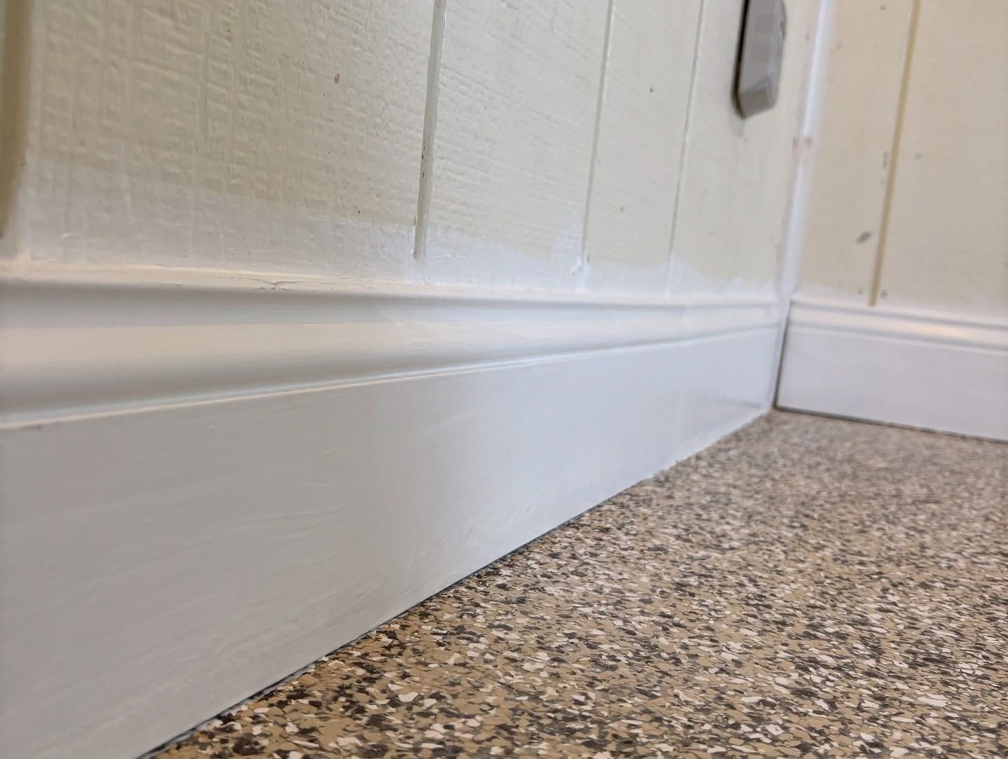 White painted baseboards and wall paneling in a room with a speckled floor.