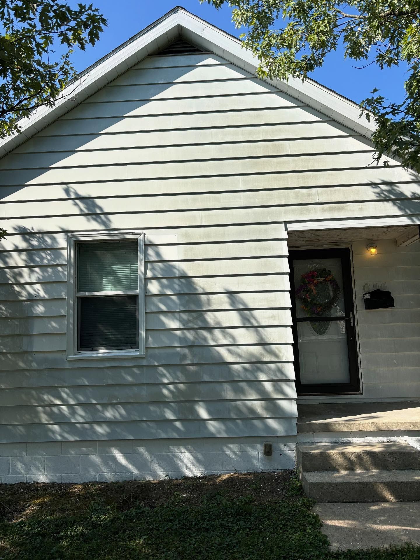 Small, light-colored house with a single window and doorway. Gray siding and a small porch with steps.