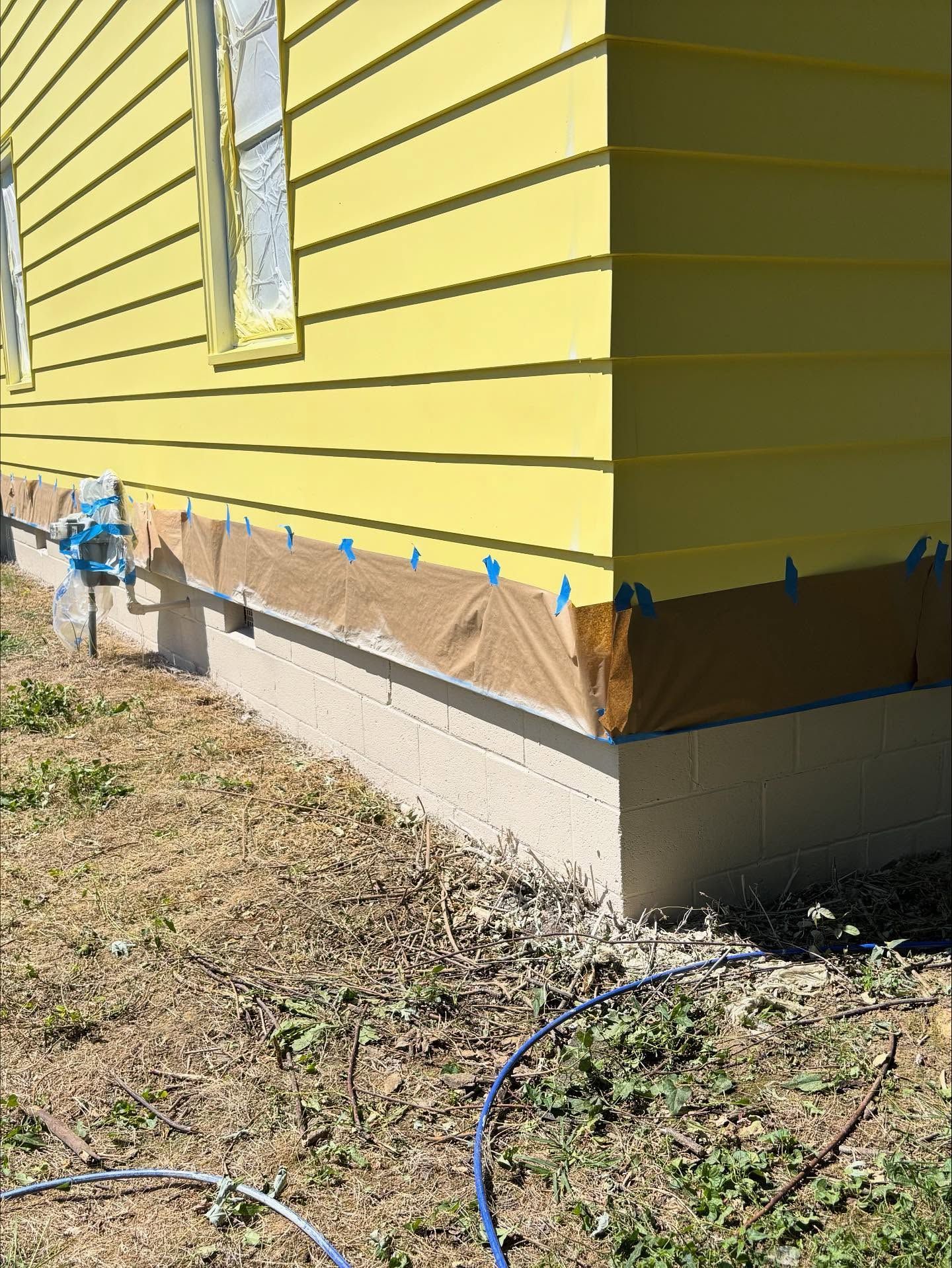 Yellow house exterior corner with masking, and tan paper covering the cinder block foundation.