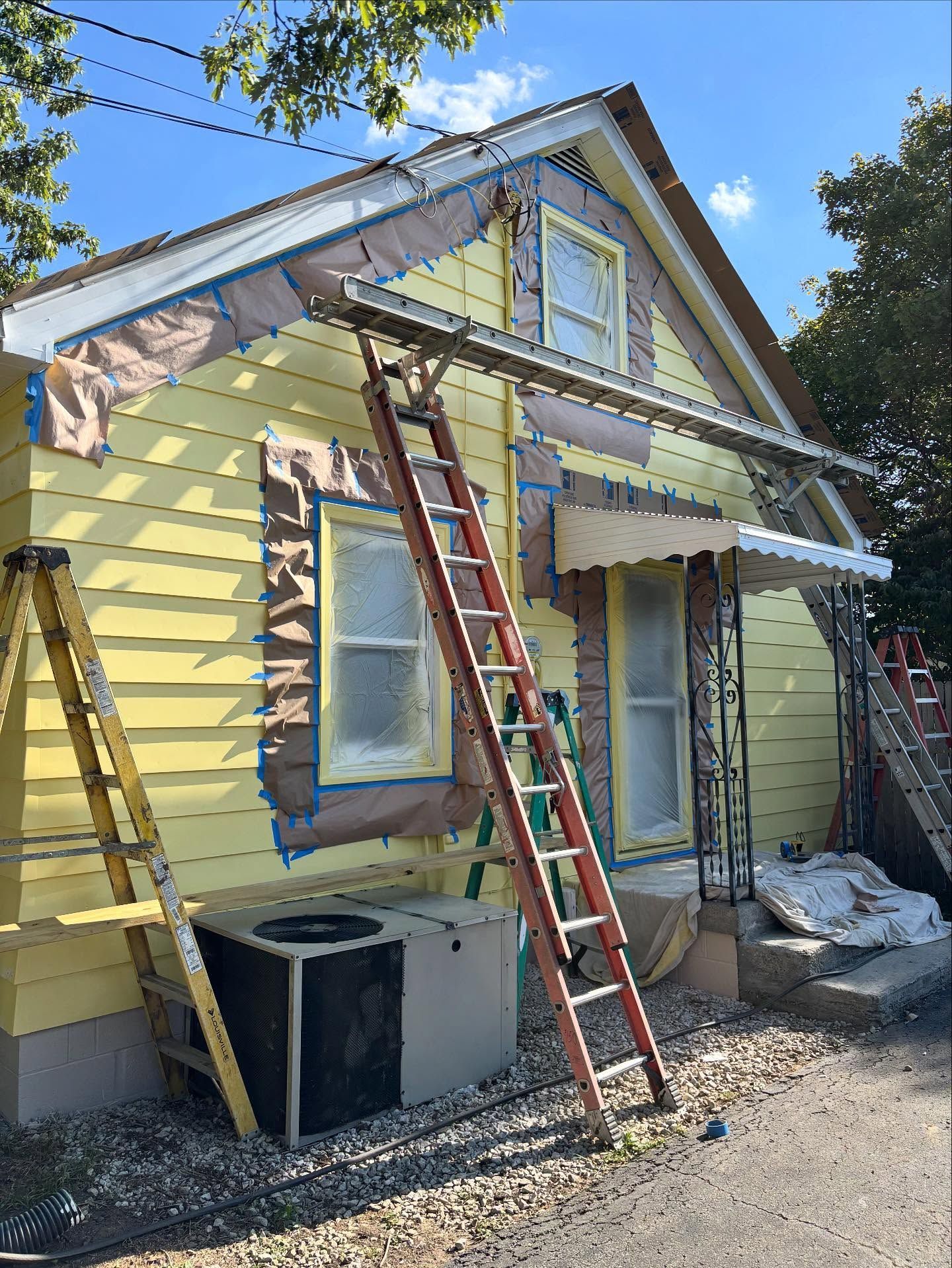 House exterior being painted yellow with ladders and protective tape on windows and trim.