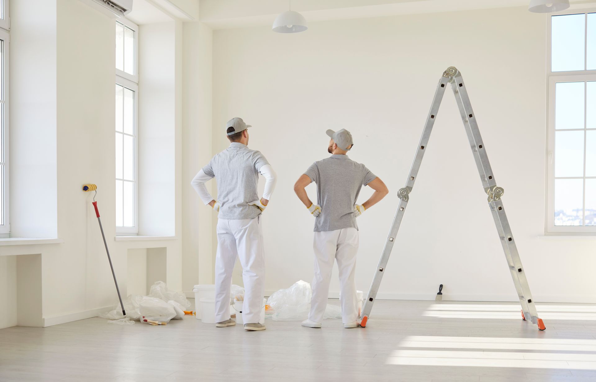 Two painters in white overalls and hats stand looking at a wall in a bright room with a ladder and supplies.
