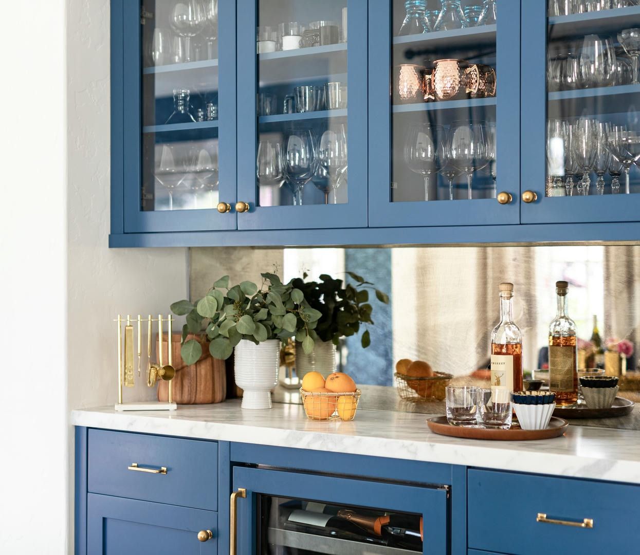 Blue home bar with glass-front cabinets holding glassware. Marble countertop with decorative items and built-in wine fridge.