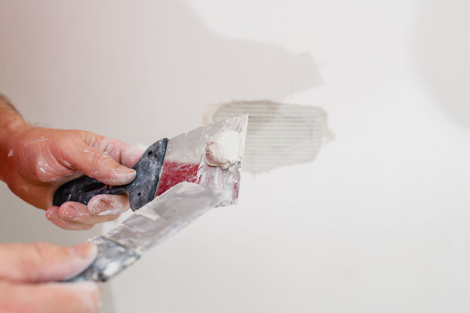 A person's hands using a putty knife to apply spackle to a wall, smoothing the surface.