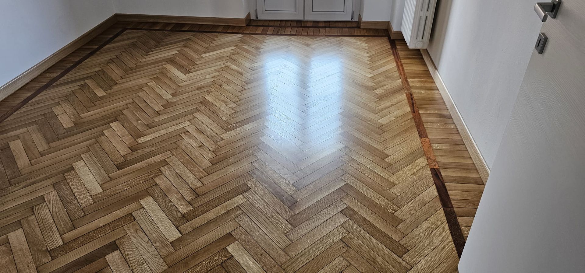 Pavimento in parquet di legno con motivo a spina di pesce, bordato e con riflessione della luce.