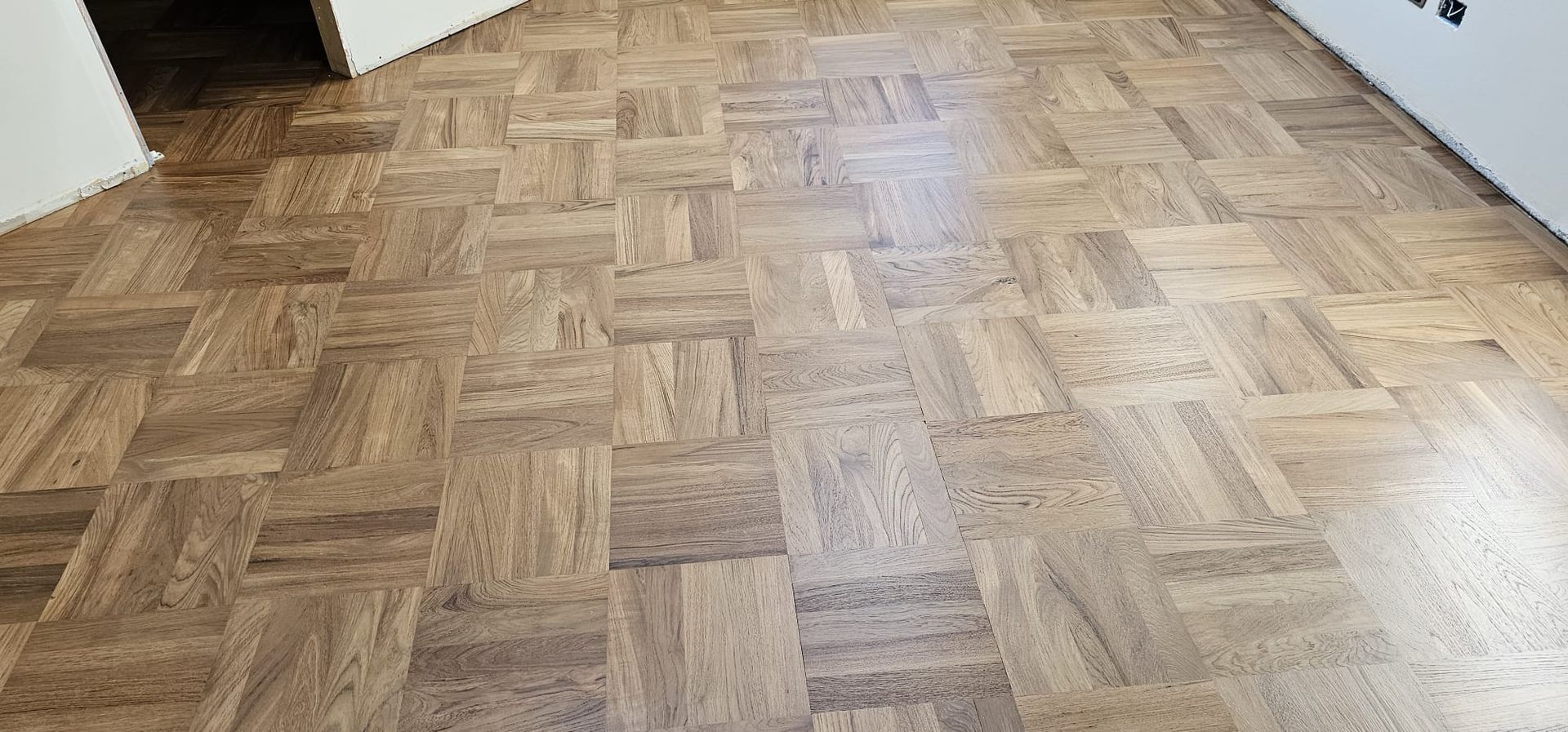 Pavimento in parquet di legno, marrone chiaro, in una stanza, con una parete bianca nell'angolo in alto a sinistra.