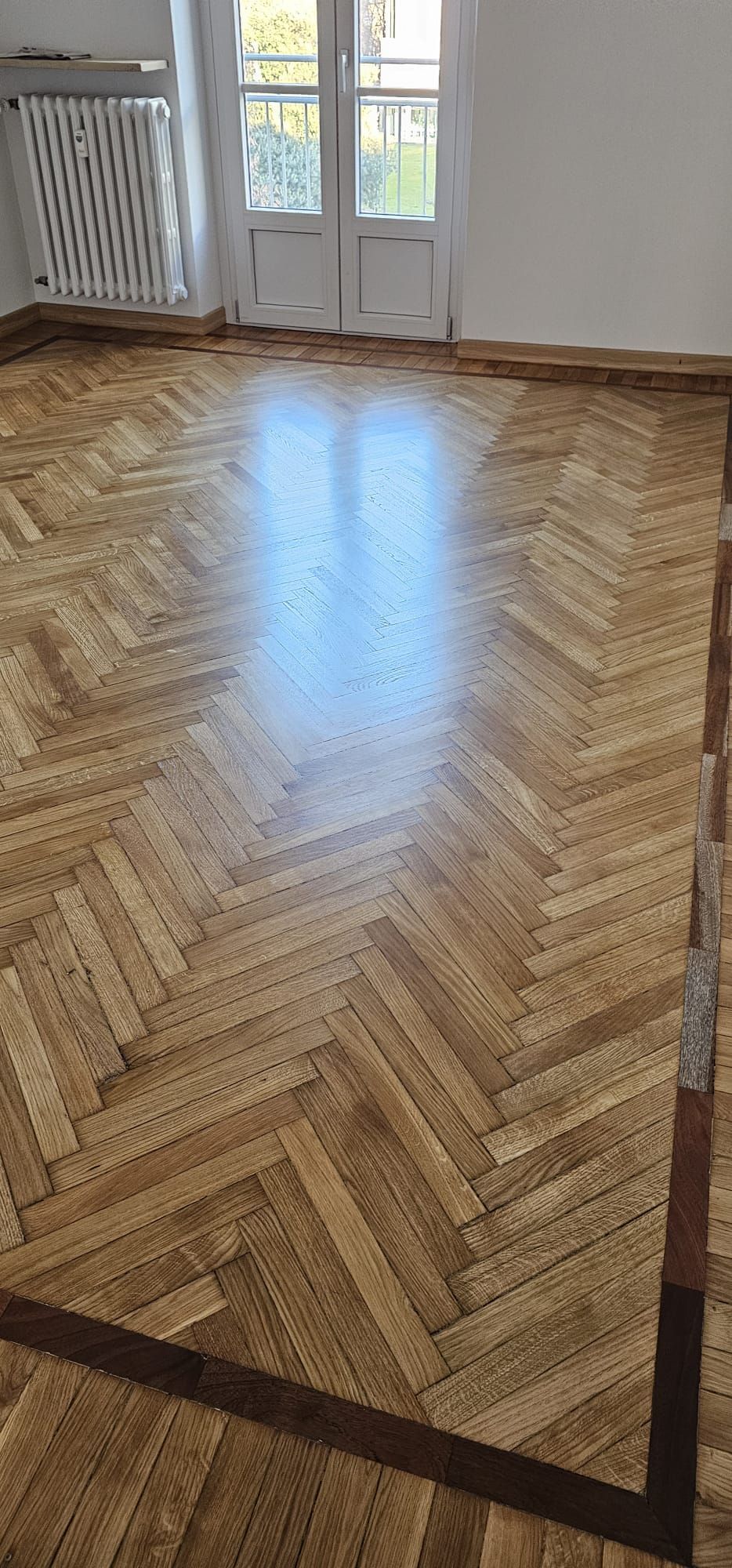 Pavimento in parquet a spina di pesce, che riflette la luce. Bordato con una finitura in legno più scuro.