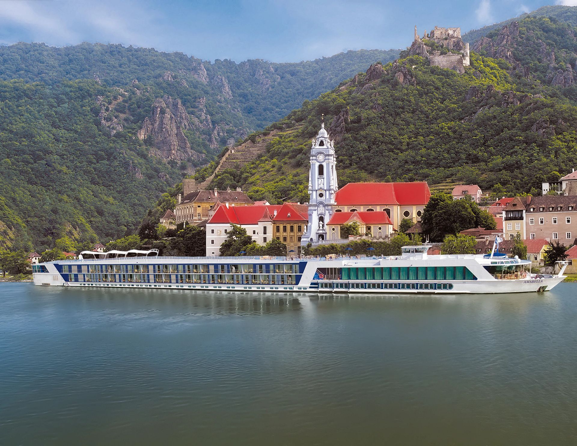 River cruise ship docked in front of a town with a church and castle, against a backdrop of green mountains.
