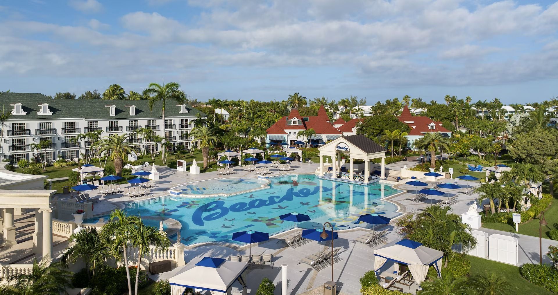 Aerial view of a Beaches resort pool area with buildings, trees, and blue water under a partly cloudy sky.