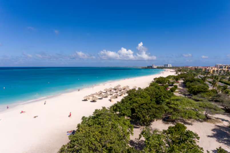 White sandy beach with turquoise water, palm umbrellas, and green trees under a blue sky.