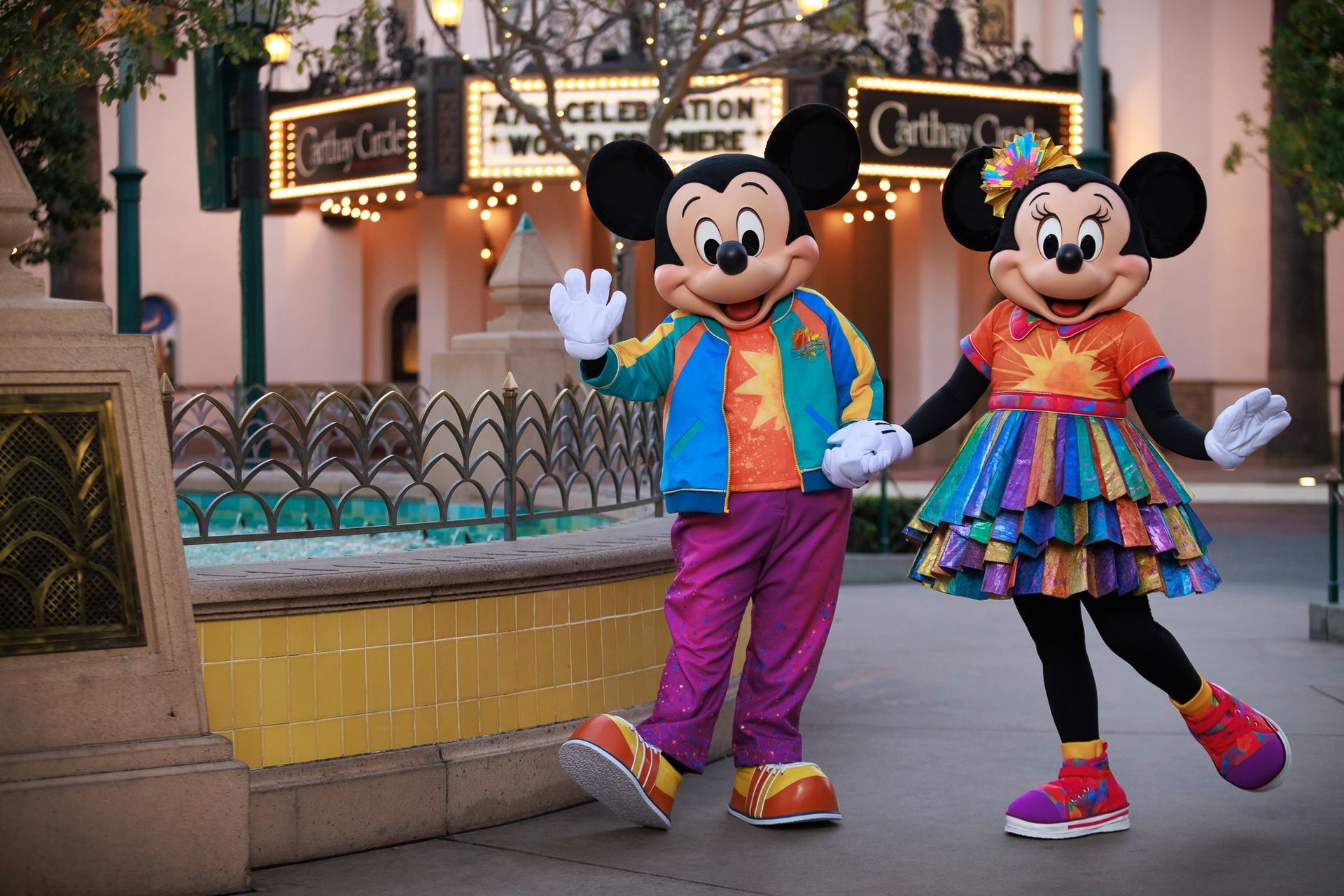 Mickey and Minnie Mouse in colorful outfits waving in front of the Carthay Circle Theatre at Disneyland.
