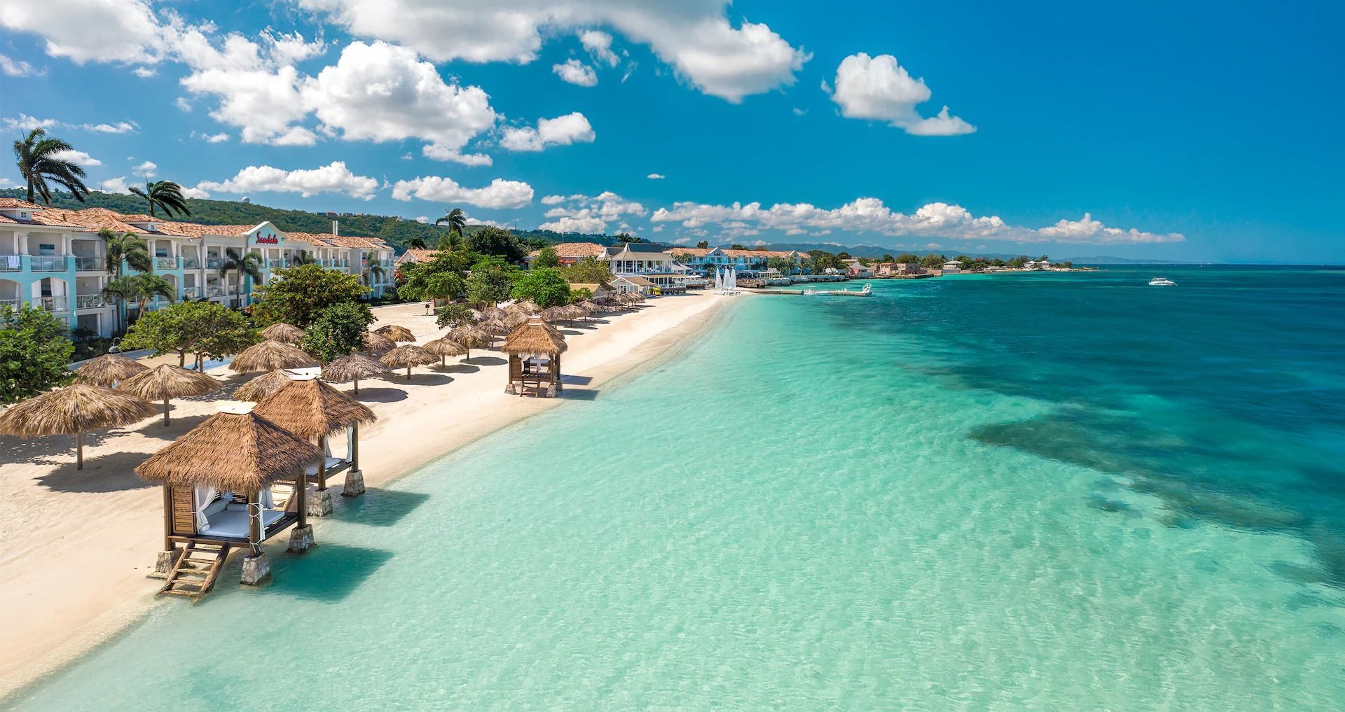 Beach with white sand, turquoise water, thatched roof cabanas, resort buildings, and a clear blue sky.