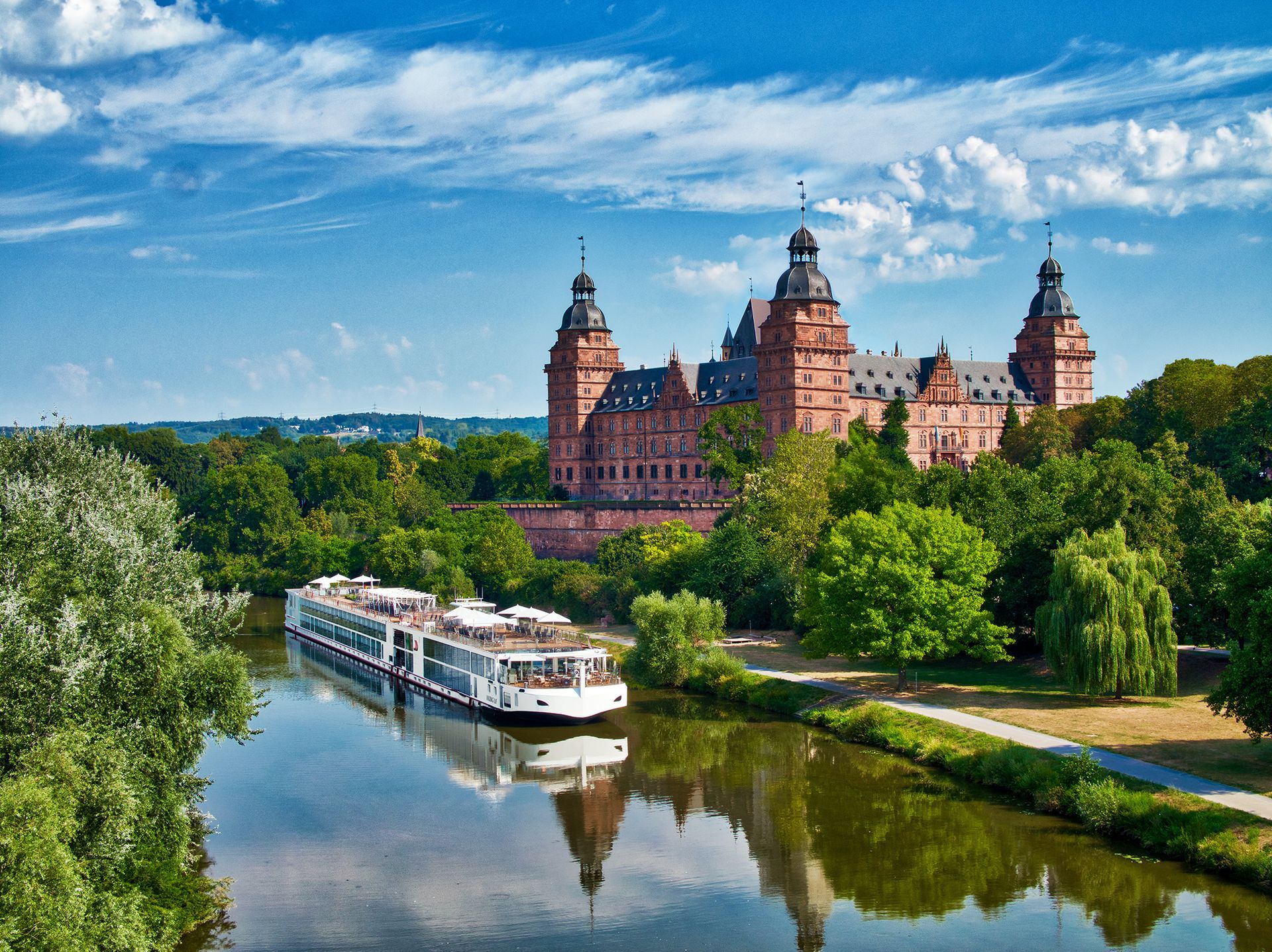 River cruise ship passing a large castle on a river, under a blue sky.