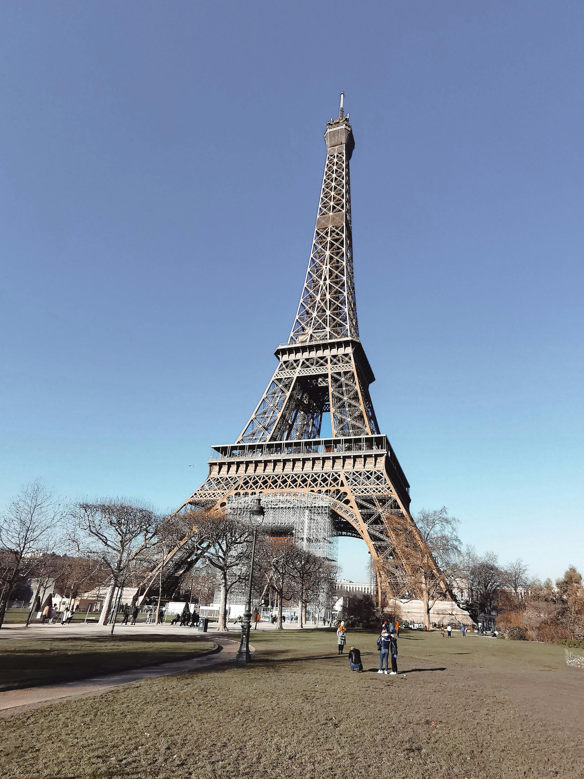 Eiffel Tower against a clear blue sky, seen from a grassy park. People are walking nearby.