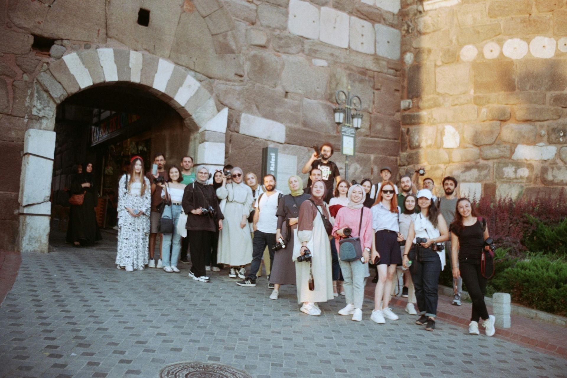 Group of people standing in front of a stone archway and wall; cobblestone ground; sunny day.