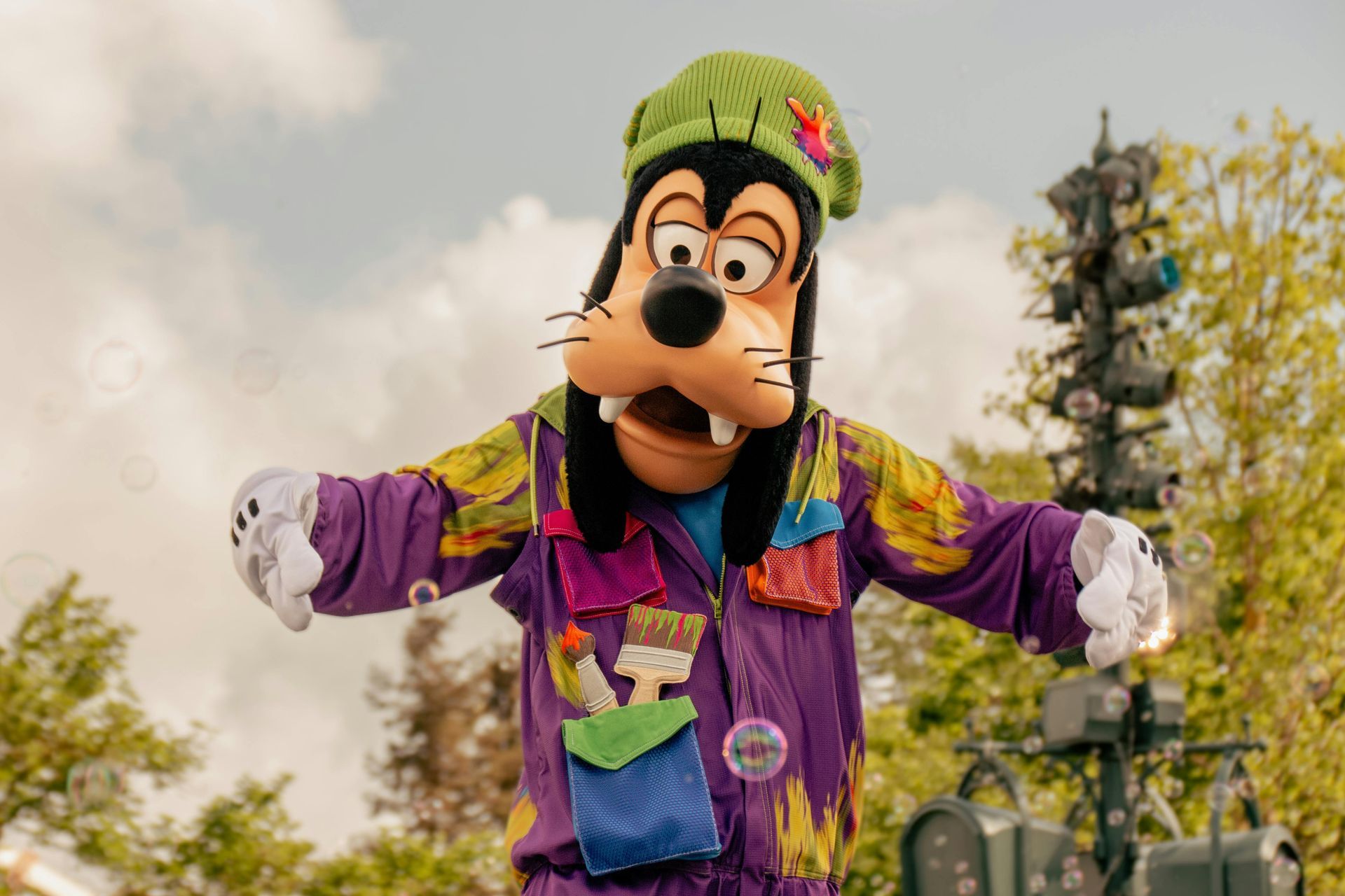 Goofy in purple paint-splattered overalls and green hat, arms outstretched, open-mouthed, outdoors.