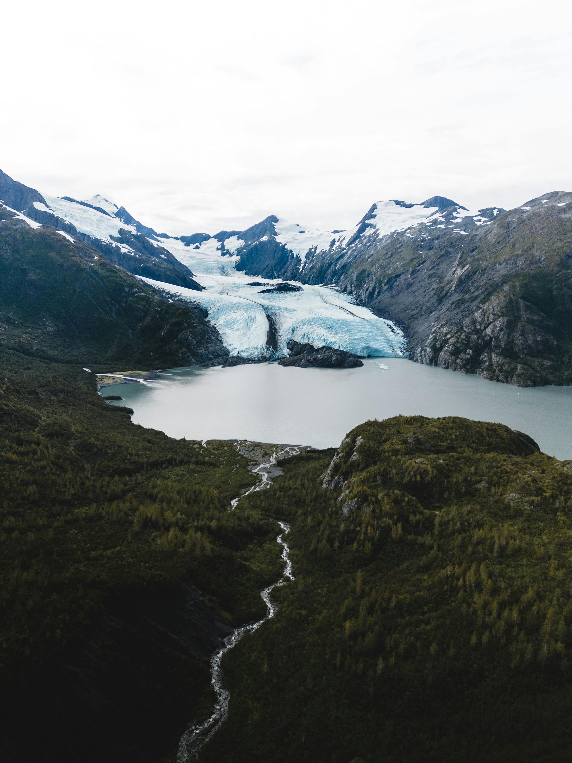 Glacier and lake surrounded by forested mountains under a cloudy sky.