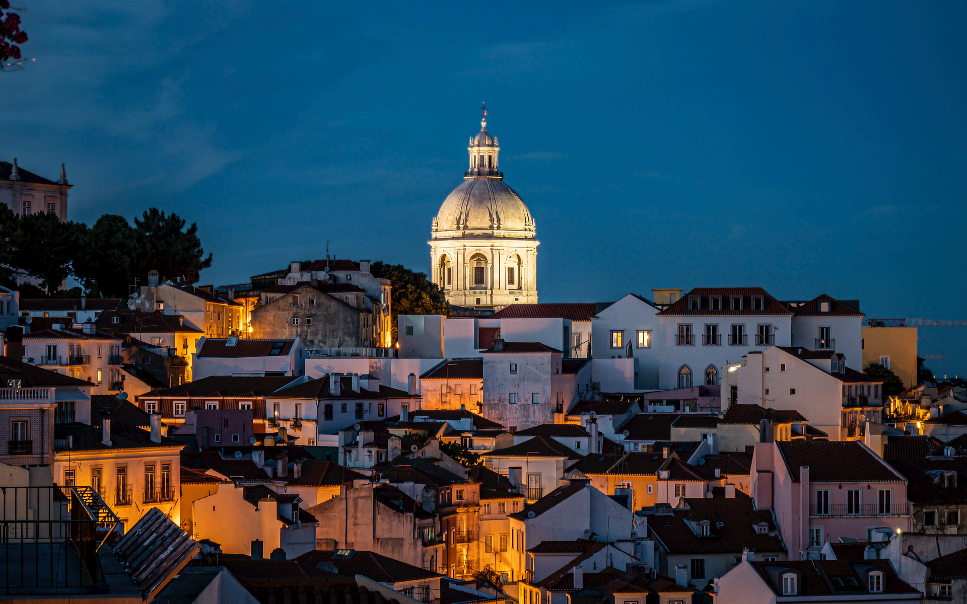Cityscape at dusk, illuminated buildings with a domed church in the center against a deep blue sky.