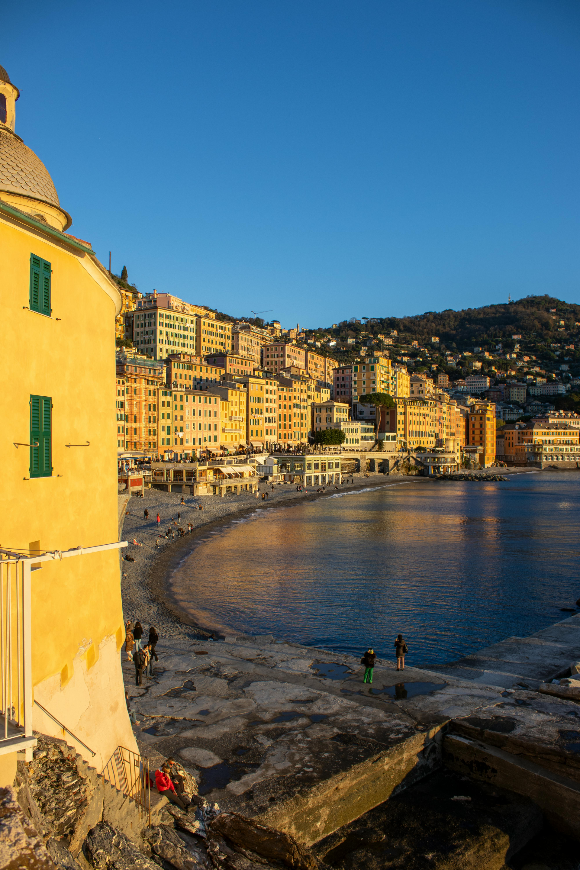 Coastal Italian town at sunset. Yellow building in foreground, colorful buildings on hillside. Blue sky and water.
