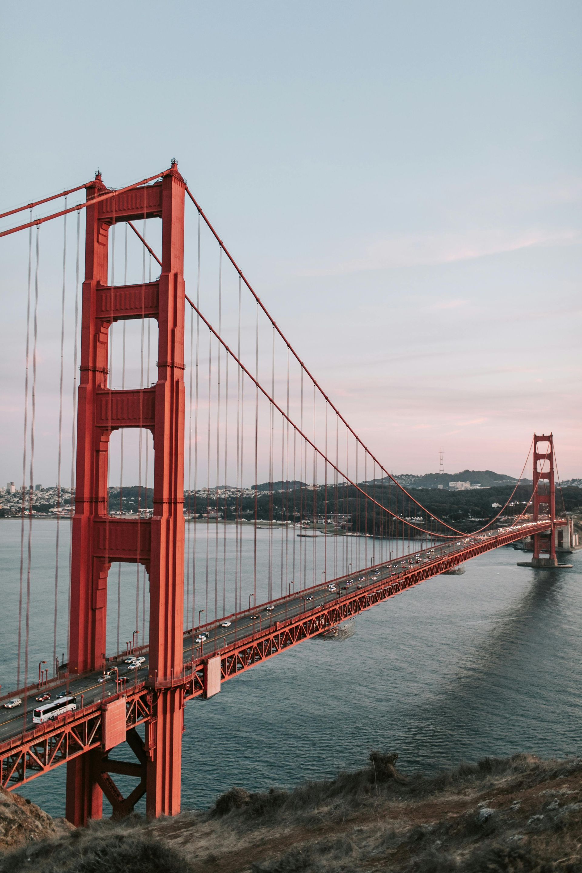 Golden Gate Bridge spans across water, cars travel, red and orange towers against pastel sky.