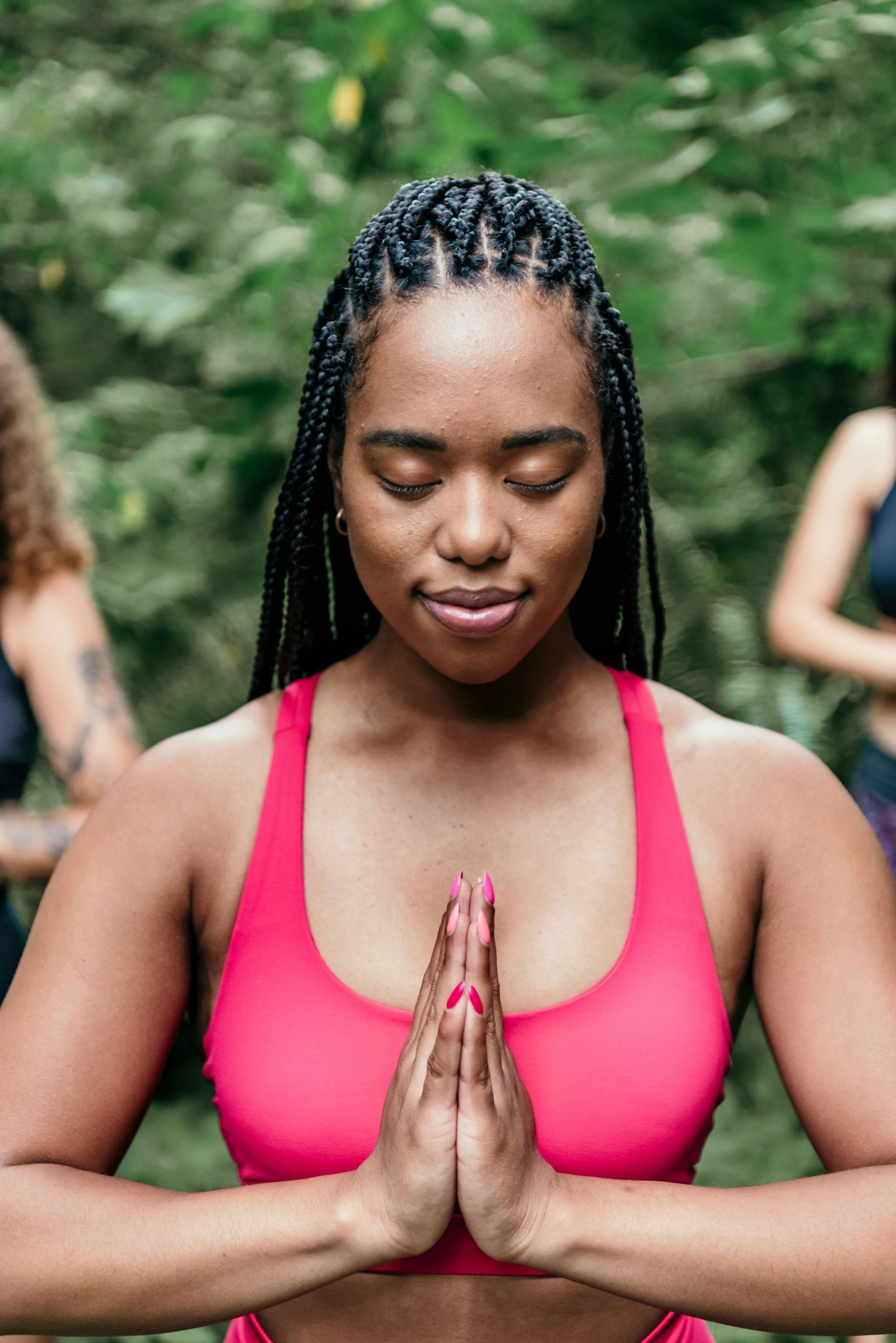 Woman in pink sports bra with hands in prayer position, eyes closed, surrounded by greenery.