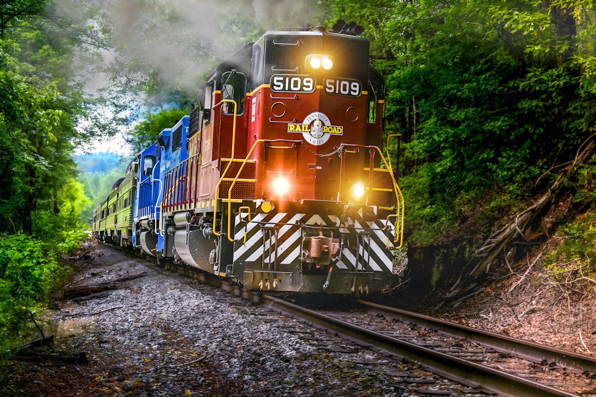 A red and blue train locomotive pulling passenger cars through a lush, green forest on a sunny day.