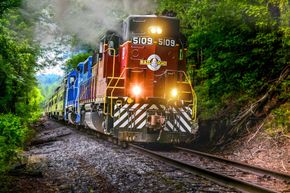 A red and blue train locomotive pulling passenger cars through a lush, green forest on a sunny day.
