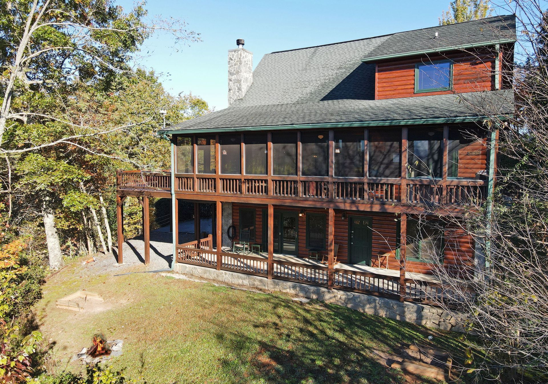 A two-story brown wood cabin with a large screened-in porch and stone chimney, nestled among trees on a sunny hillside.