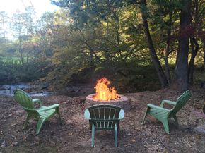 Three green Adirondack chairs arranged around a stone fire pit with a bright flame, set in a wooded area by a creek.