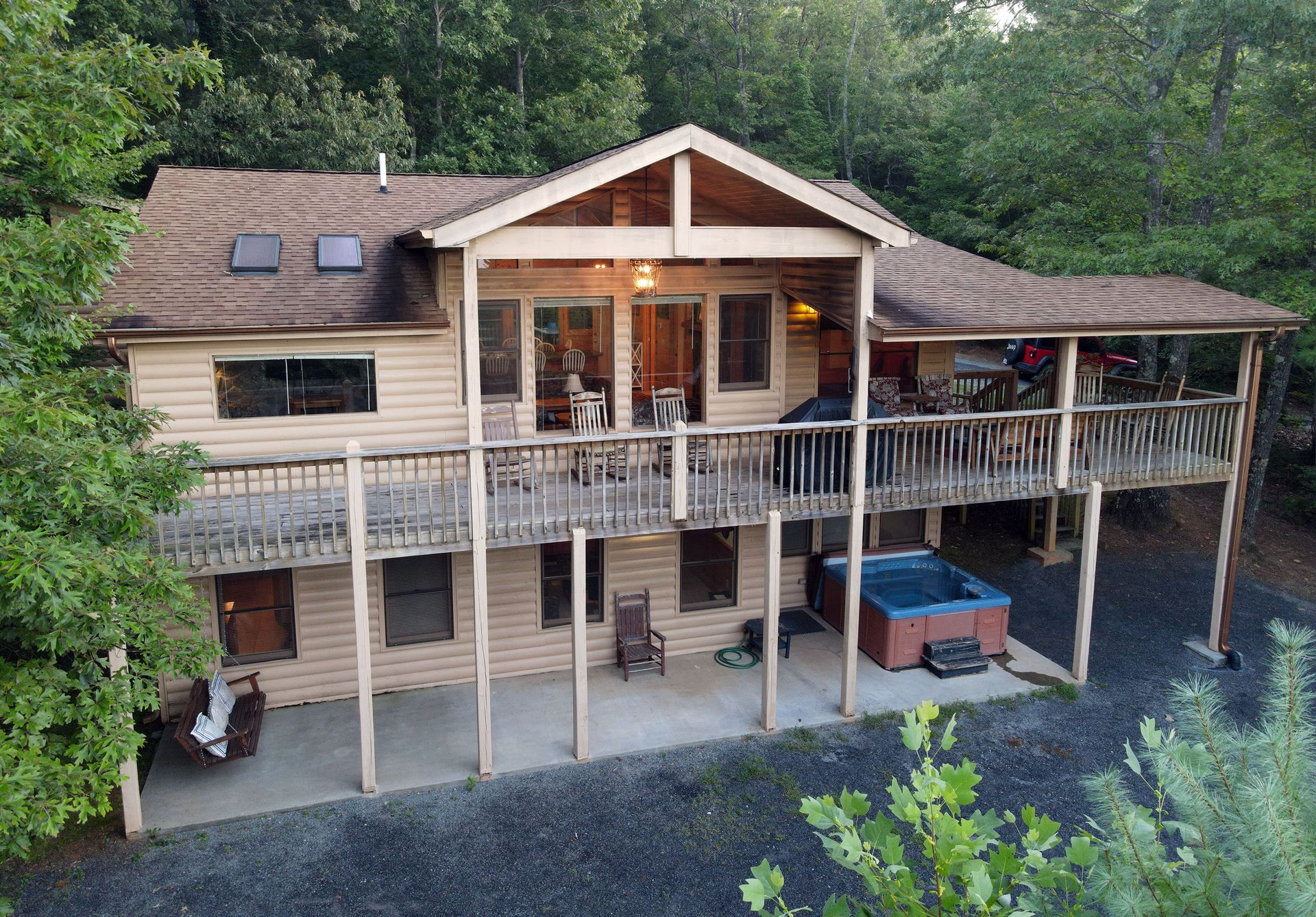 A two-story cabin with a wooden exterior, a covered wrap-around deck, and a lower-level patio with a hot tub, set in woods.