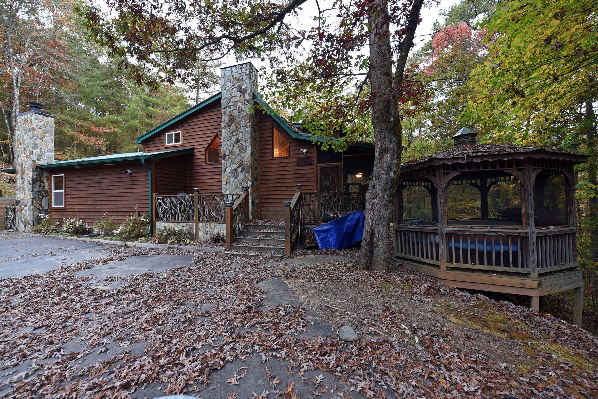 A wood cabin with a stone chimney and a covered gazebo, surrounded by autumn trees and fallen leaves.