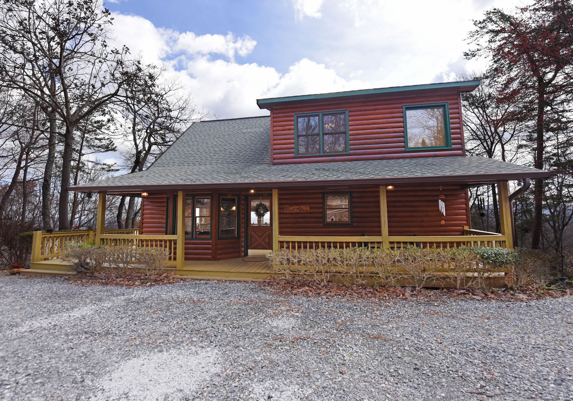 A two-story red log cabin with a wide, covered front porch and a gravel driveway, surrounded by trees under a cloudy sky.