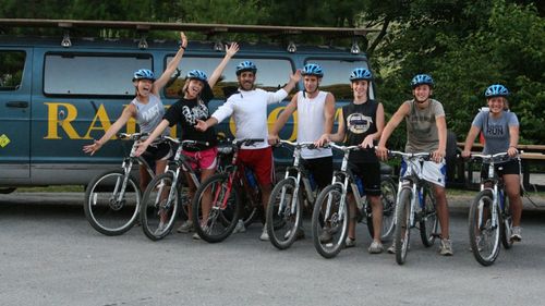 A group of eight cyclists with helmets pose with their mountain bikes in front of a dark blue van outdoors.