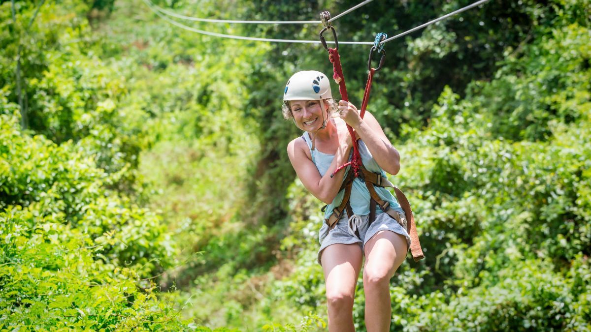 A smiling person wearing a helmet and harness glides along a zip line above a lush, green forest canopy.