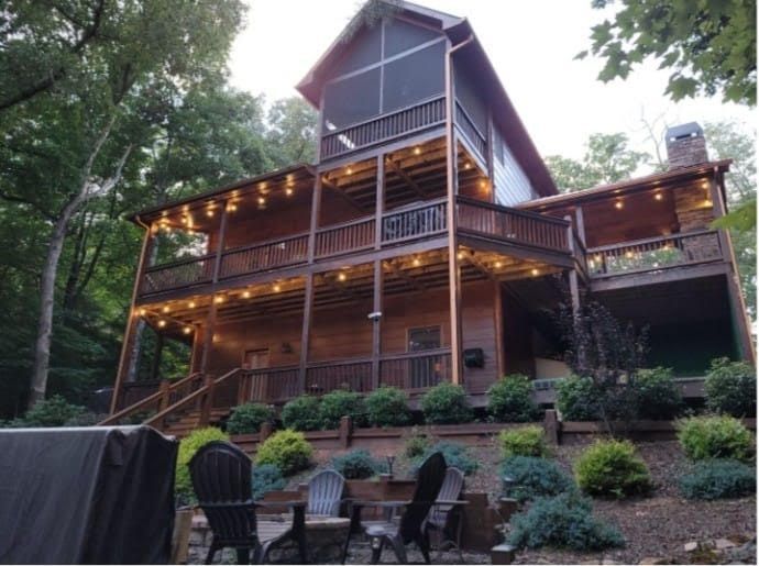 A multi-story wooden cabin at twilight, featuring illuminated decks, a screened-in porch, and a patio with chairs.