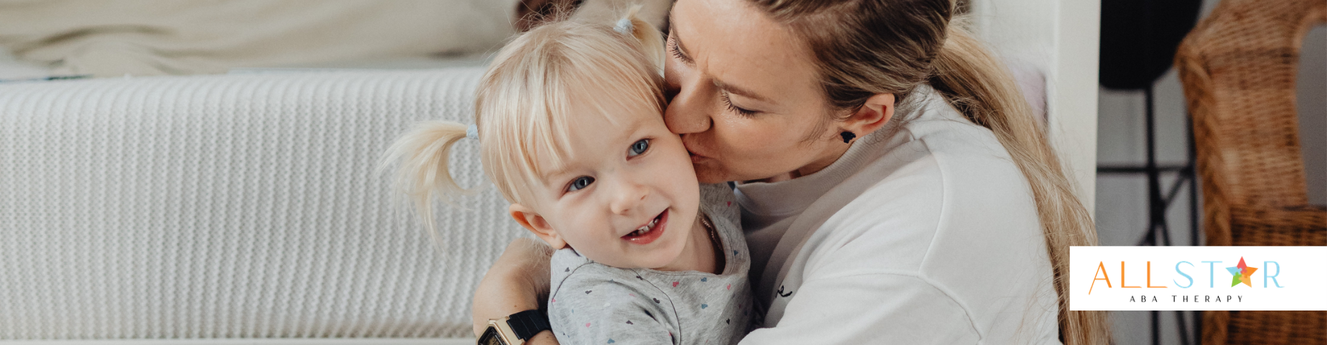Woman kissing a smiling child with blonde pigtails on the cheek; sitting indoors near a textured wall.
