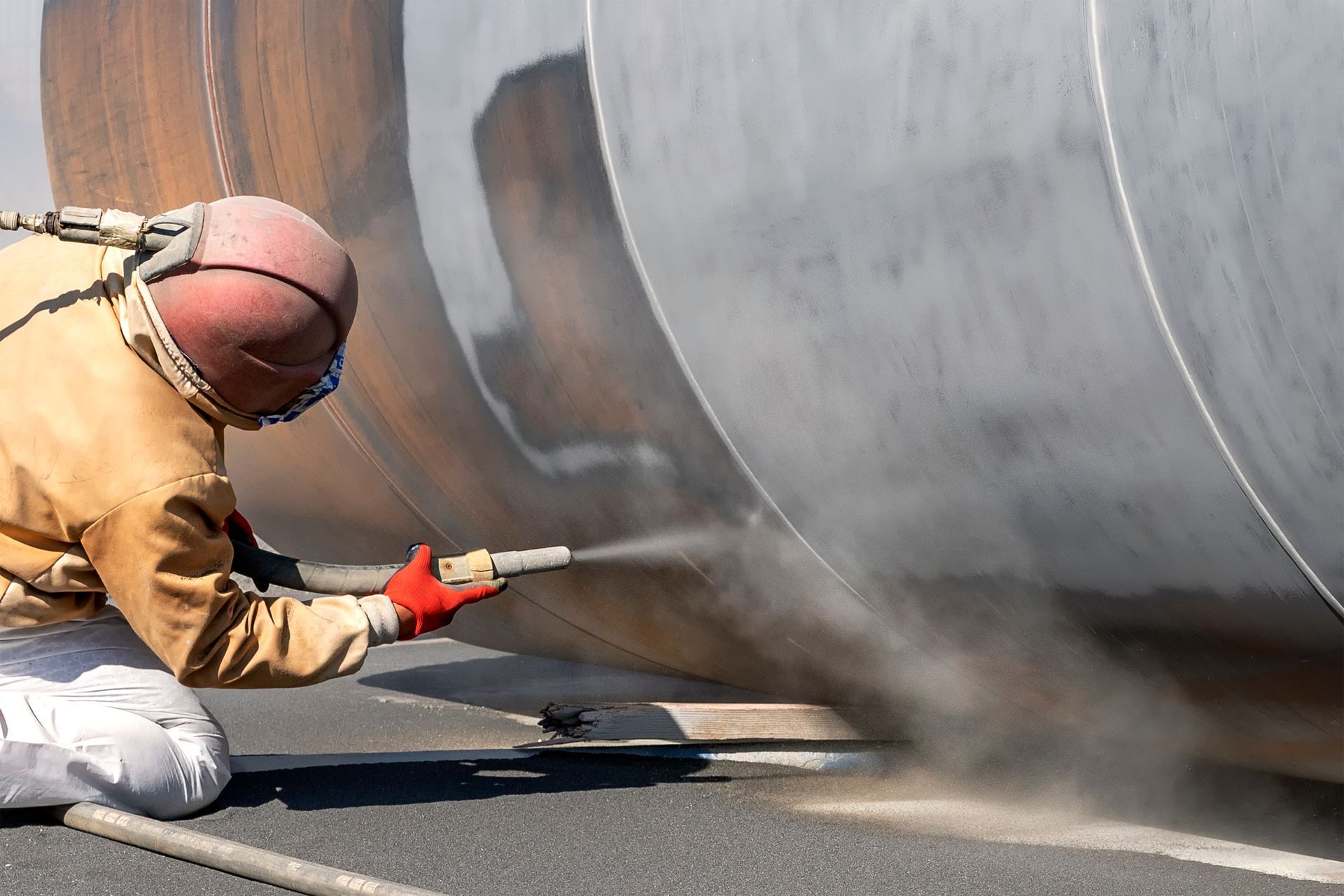 Worker in protective gear sandblasting a large metal tank with a high-pressure hose.