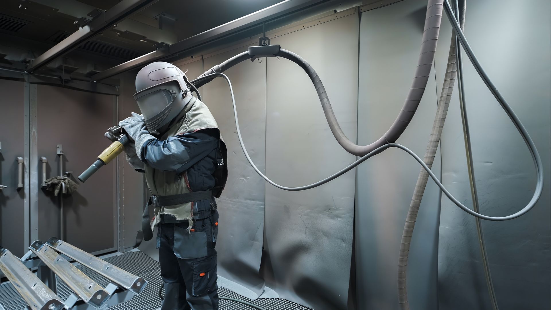 Worker in protective gear using an abrasive blasting hose inside a blast booth.