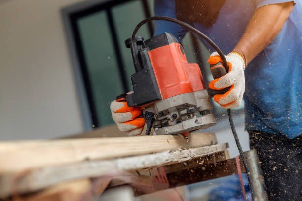 Worker using electric router to shape wood board with flying sawdust.