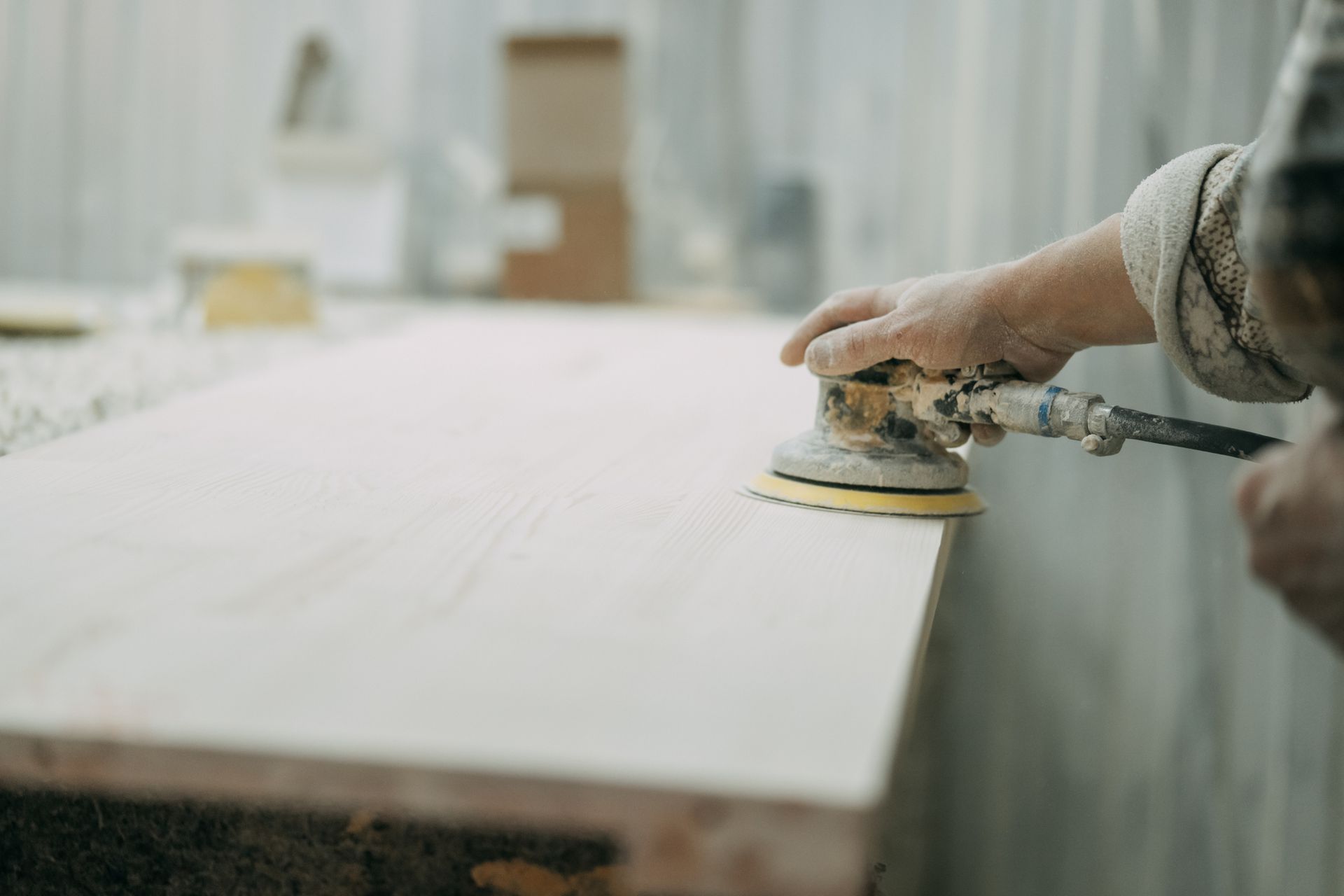 Carpenter is using an orbital sander to smooth a wooden plank in a workshop.