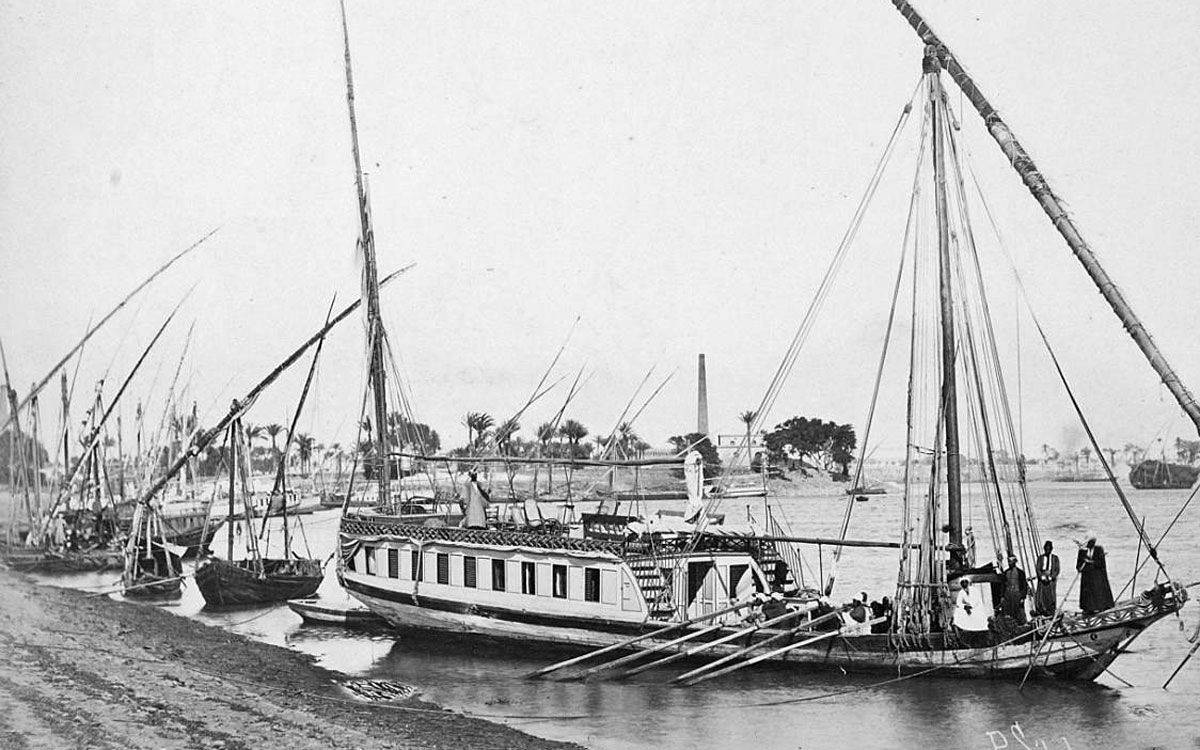 A historical photo of a Dahabiya Kingfisher sailing on the Nile in Egypt in 1899. The photo is in black and white and shows the boat from a side view. The Dahabiya Kingfisher is a traditional wooden boat that can be rented for private cruises between Luxor and Aswan.