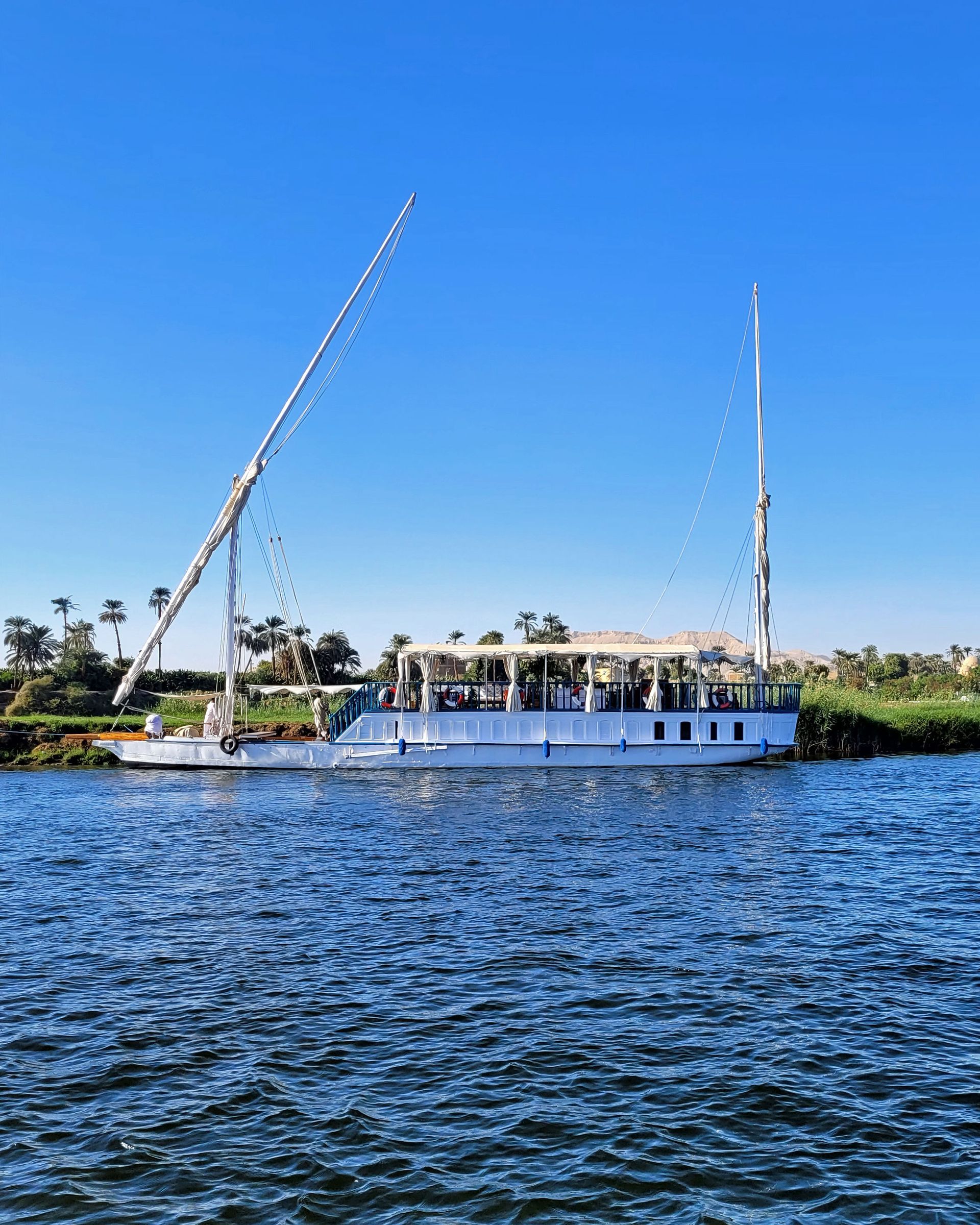 In this photo, you can see the Dahabiya Kingfisher, a traditional wooden boat that dates back to 1899. The boat has a graceful white hull, it sails along the Nile river, the longest and most ancient river in the world. The boat offers private cruises for travelers who want to experience the beauty and culture of the Nile valley, where they can visit temples, tombs, and villages along the way.