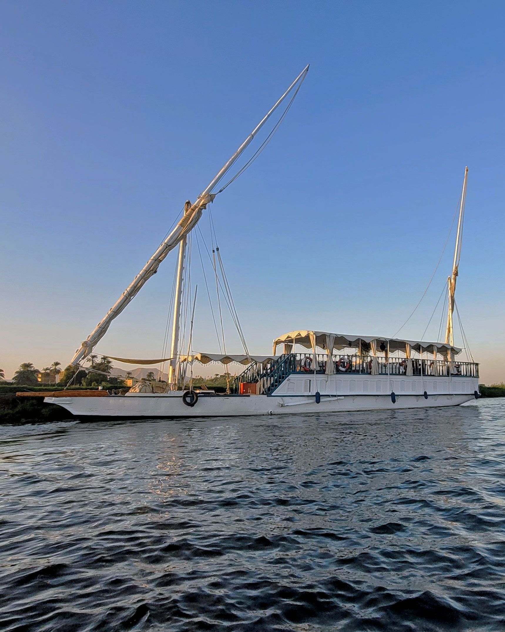 A picture of a traditional sailing boat on the Nile river, with a blue sky and green palm trees in the background. The boat is called the Dahabiya Kingfisher and it provides private cruises between Luxor and Aswan in Egypt, following the route of the original 1899 vessel.